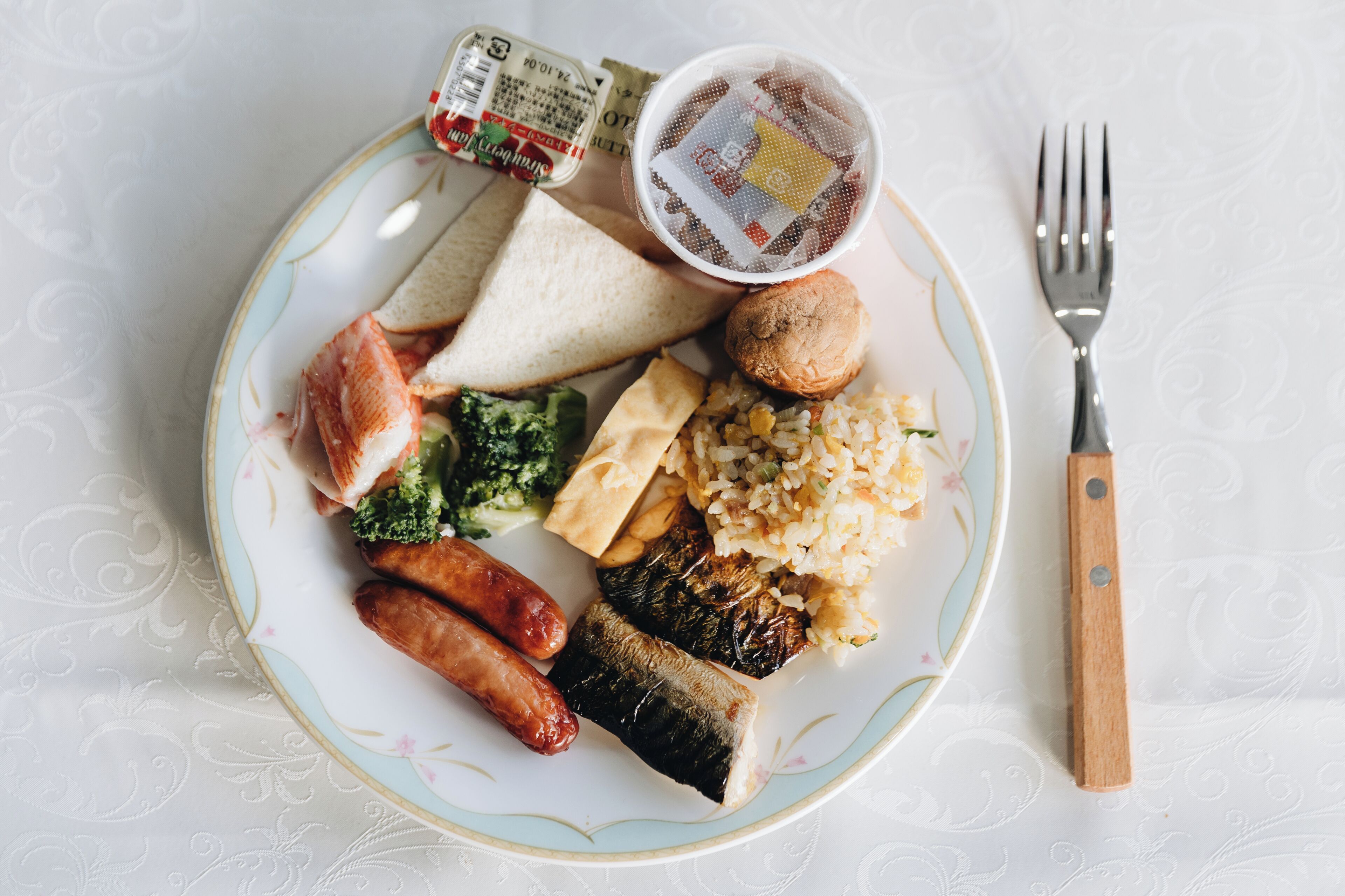 Un plato con diversos alimentos, incluyendo pan, salchichas, champiñones y arroz, acompañado de una taza de té y un tenedor sobre una mesa con mantel estampado.