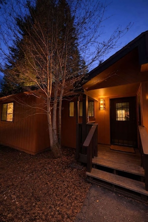 A wooden cabin with a porch, a tree in front, and a lit exterior. - Cozy Shaver Lake Studio with Pool and Hot Tub, In Bretz Mill Condominiums! (Shaver Lake)