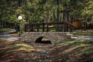 Un puente de piedra con pasarela de madera sobre un arroyo, rodeado de árboles y un farol.
