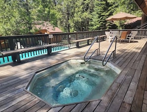 A wooden deck with a hot tub, outdoor seating, and a view of trees. - Cozy Shaver Lake Studio with Pool and Hot Tub, In Bretz Mill Condominiums! (Shaver Lake)