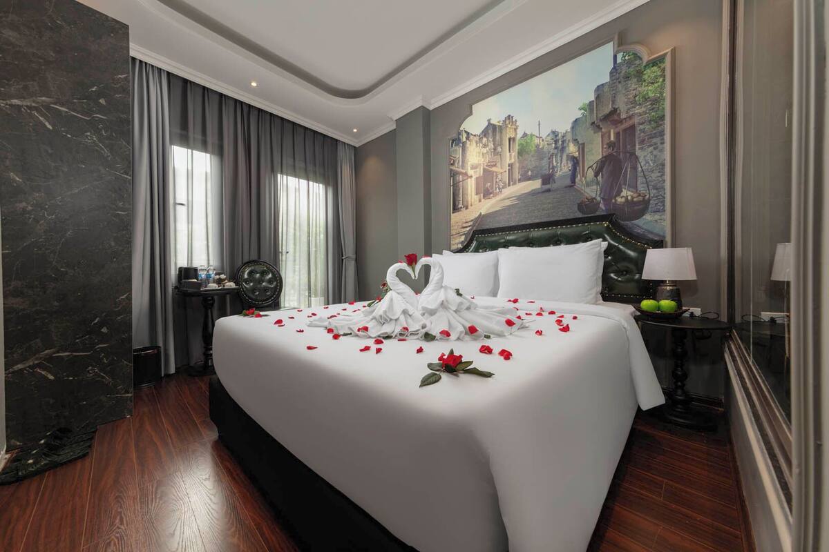 Habitación de hotel con una cama grande, un cuadro de una escena callejera, un escritorio con una lámpara y una silla.