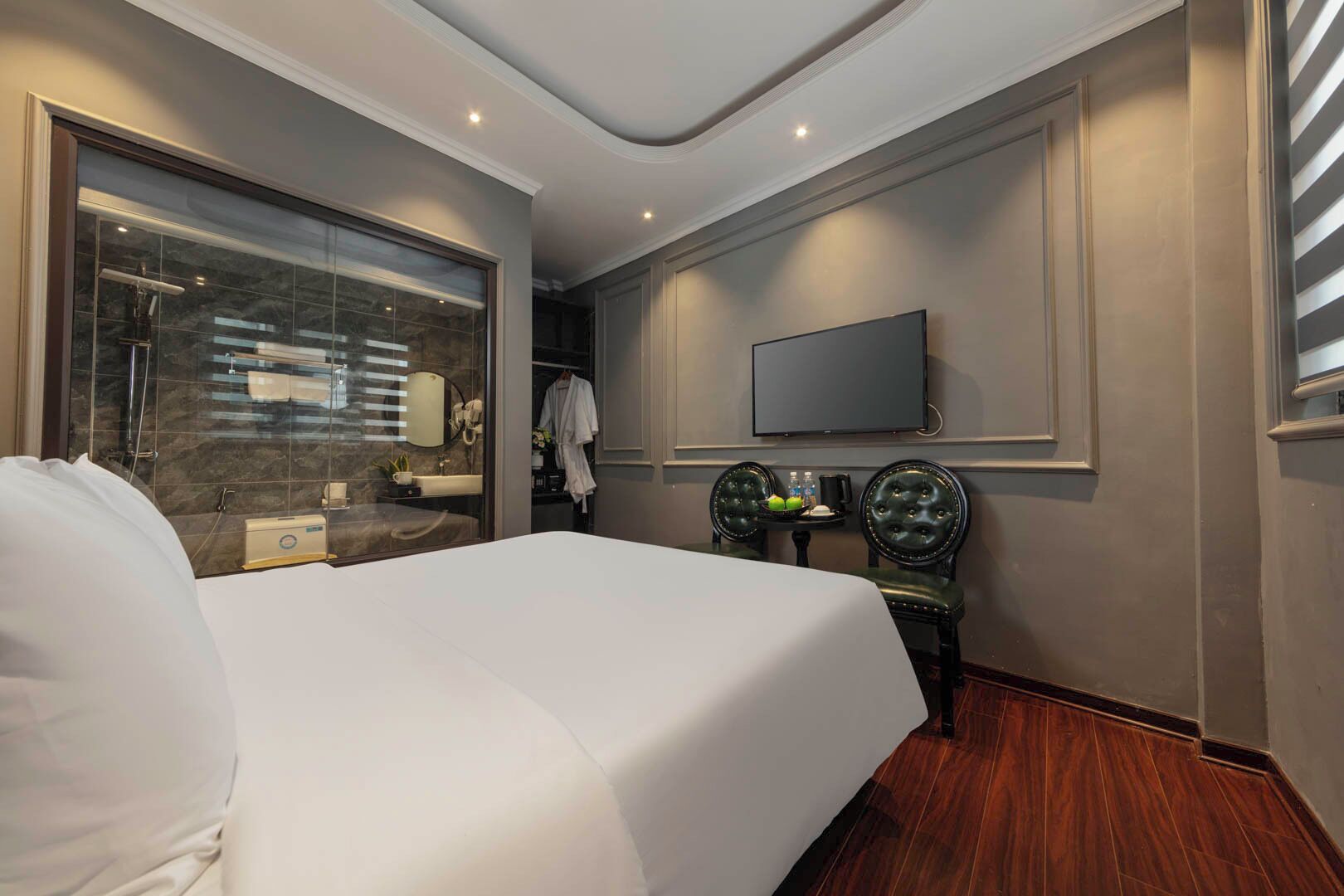 Habitación de hotel con una cama grande, un televisor montado en la pared, una mesita con dos sillas y un baño visible a través de un mamparo de vidrio.
