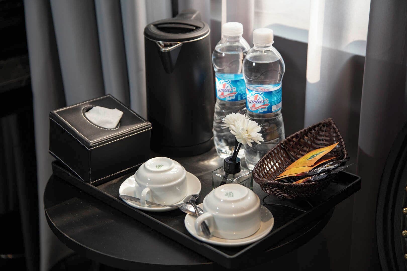 Un plateau contenant une théière, deux bouteilles d’eau, une boîte de mouchoirs, une petite tasse blanche, une soucoupe blanche, un petit vase avec une fleur blanche et un panier rempli de serviettes pliées.
