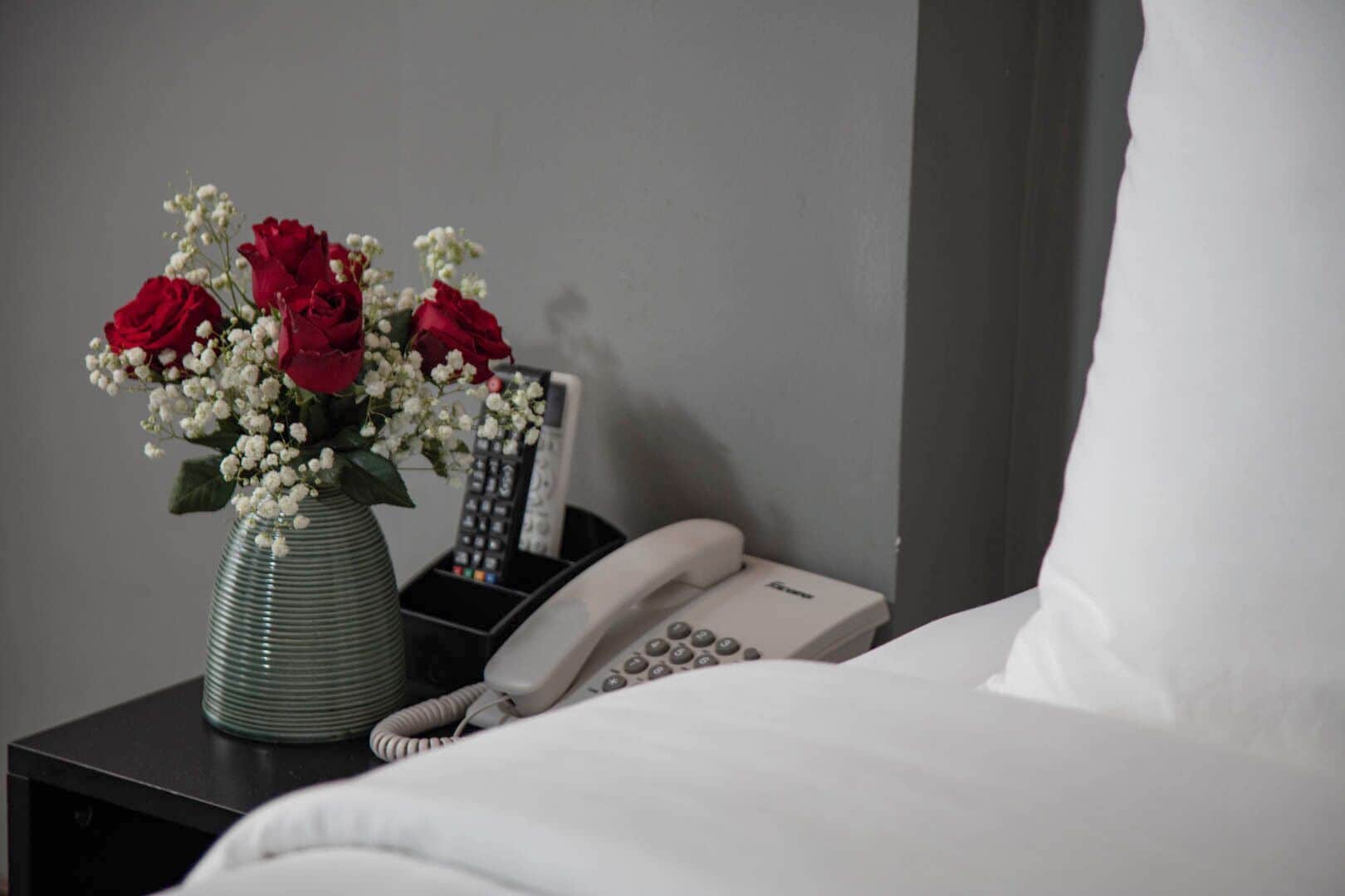 Une chambre d’hôtel avec un lit, une table de chevet ornée d’un vase contenant des roses rouges et des fleurs blanches, un téléphone et une télécommande.