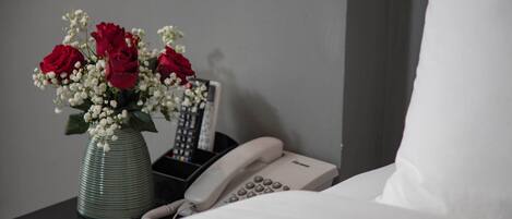 Habitación de hotel con cama, una mesita de noche con un jarrón de rosas rojas y flores blancas, un teléfono y un control remoto.