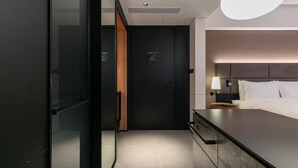 Habitación de hotel moderna con una cama grande, una mesita de noche, un televisor y una puerta que da a un pasillo.