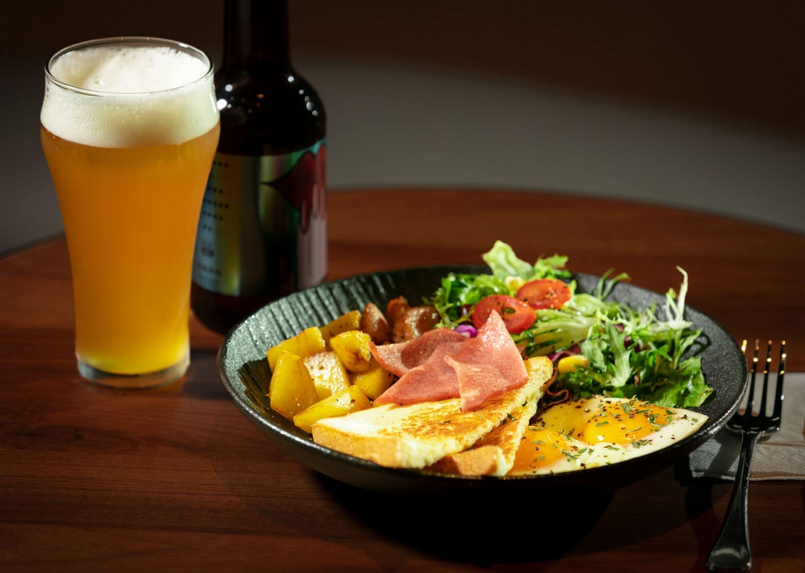 Uma refeição com torradas, ovos, presunto e salada, além de um copo de cerveja sobre a mesa.