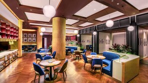Un moderno vestíbulo de hotel con suelo de madera, asientos azules, mesas redondas y una columna central.