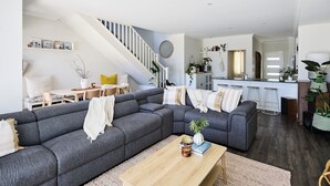 A modern living room with a grey sectional sofa, a wooden coffee table, and a staircase leading to a kitchen area. - Water’s Edge: 4BR all-ensuite. Sweeping estuary views for families and groups. (Falcon)