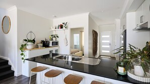 A modern kitchen with a black countertop, white bar stools, and a built-in microwave. - Water’s Edge: 4BR all-ensuite. Sweeping estuary views for families and groups. (Falcon)