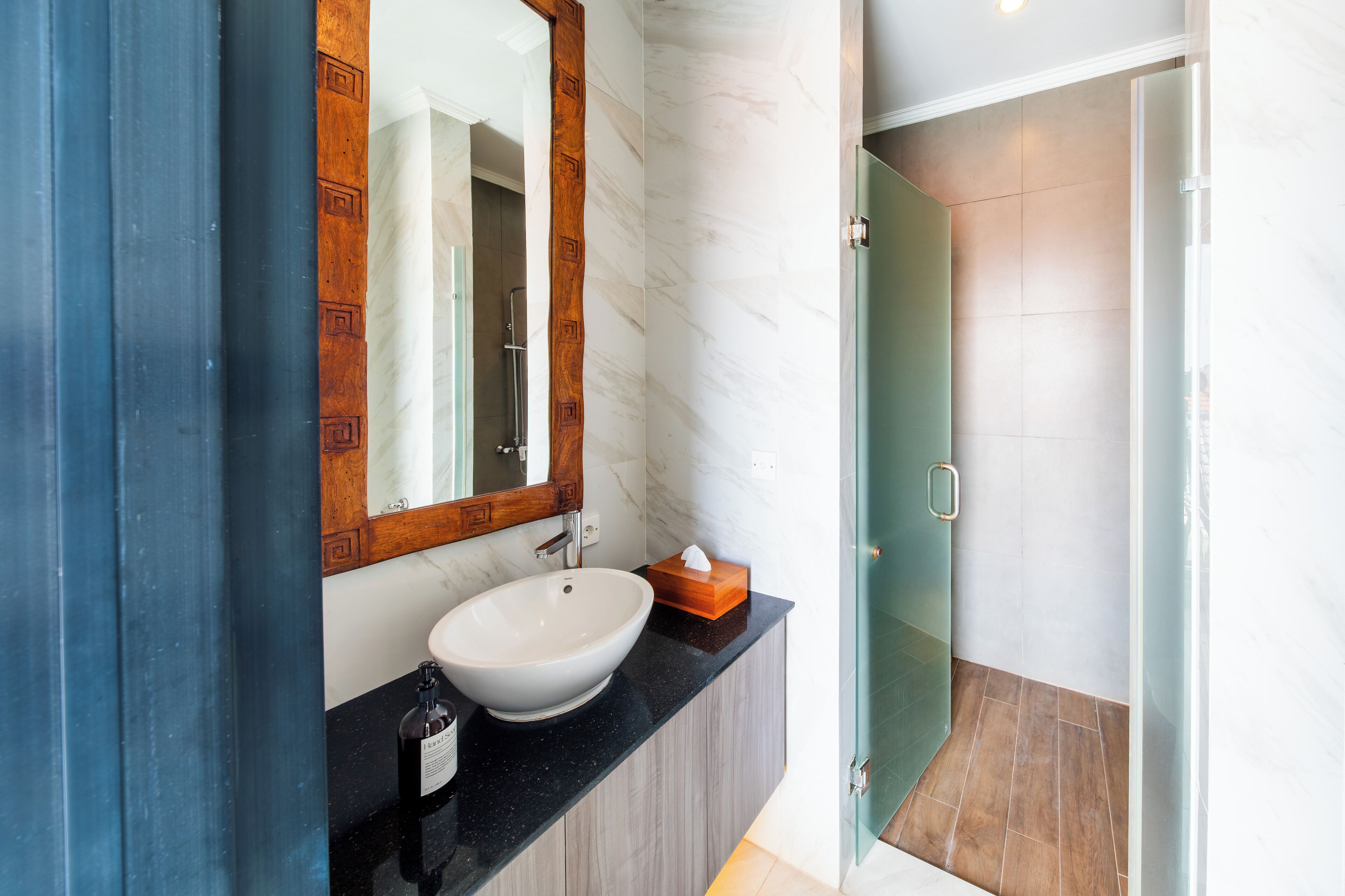 A bathroom with a white sink, a wooden-framed mirror, a glass shower enclosure, and a black countertop.