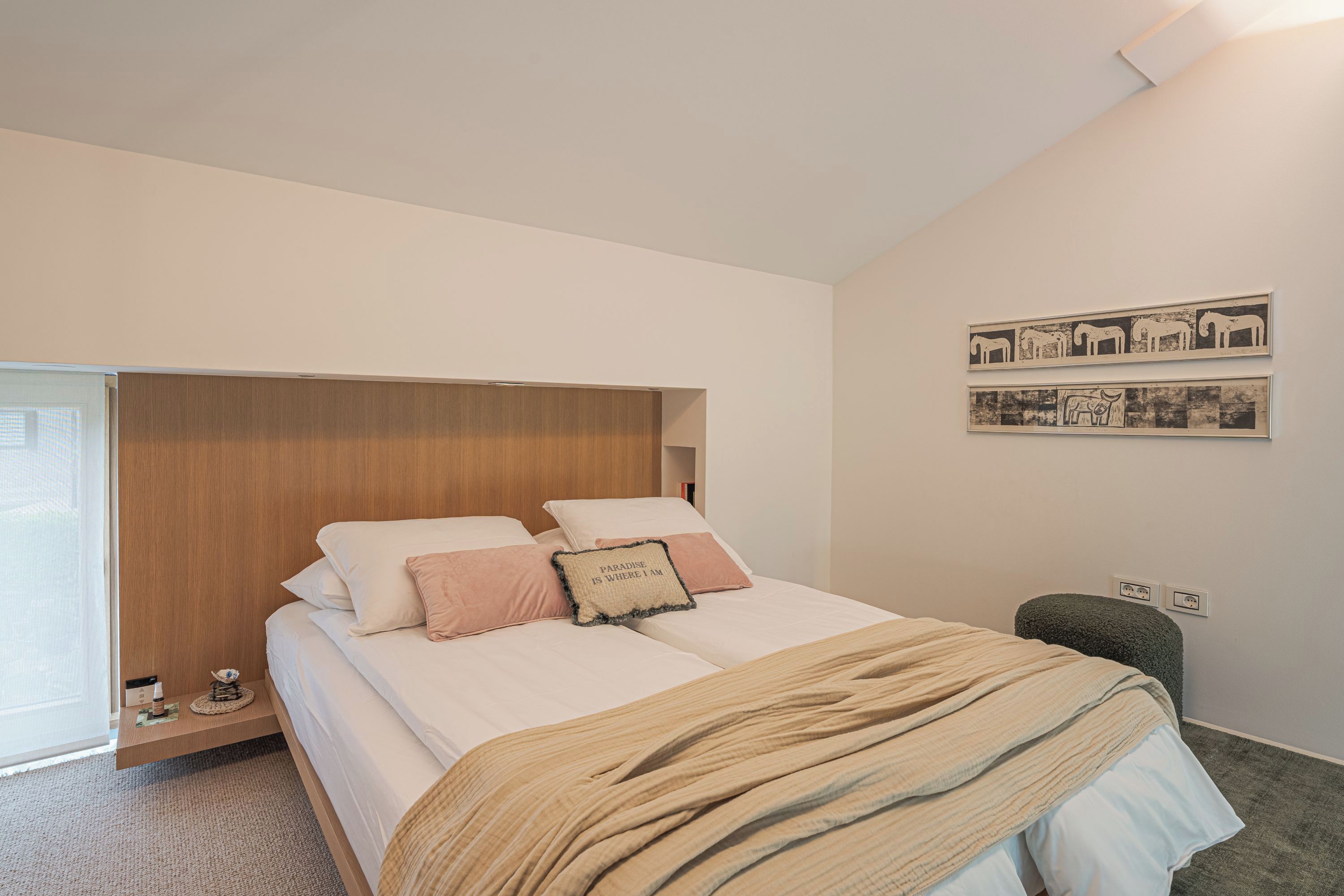 A bedroom with a bed, pillows, a blanket, a wooden headboard, a wall-mounted artwork, and a small stool.