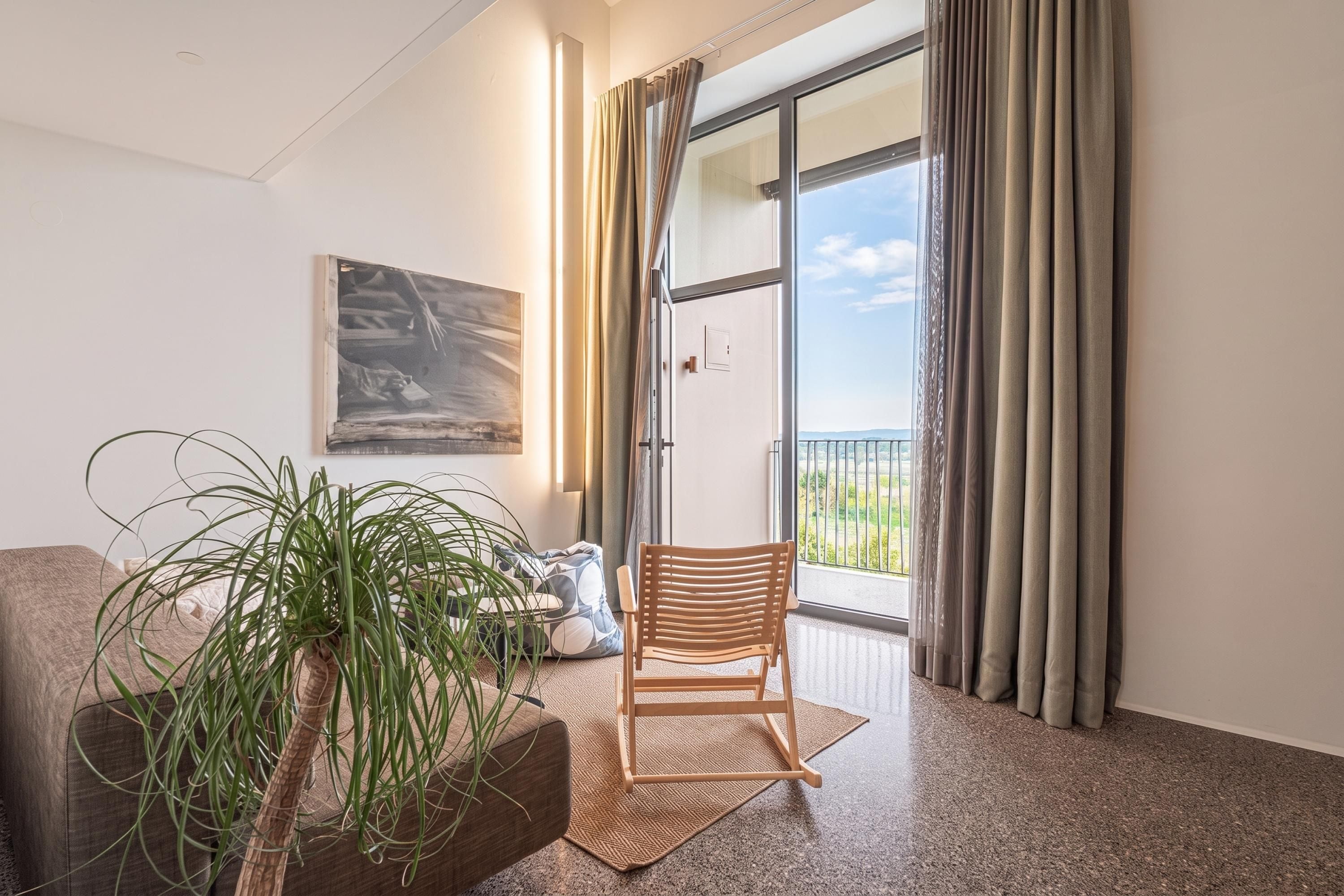 Una habitación con un ventanal grande, una silla de madera, una planta y un cuadro en la pared.