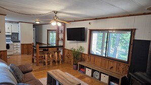 A cozy living room with a fireplace, a kitchen in the background, wooden walls, and a large window with a view of trees.