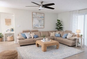 A living room with a beige sofa, a woven coffee table, a ceiling fan, a framed abstract painting, and a potted plant. - Spacious 4-bedroom house in lovely Sarasota with Private Pool (Sarasota)