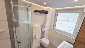 A modern bathroom with a glass-enclosed shower, a toilet, a sink, and a window with blinds. - Ferienwohnungen Luttinger am Mühlbach – Cozy Apartment (Bichlbach)