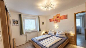A room with a bed, a window with curtains, a radiator, a ceiling light fixture, and artwork on the wall. - Ferienwohnungen Luttinger am Mühlbach – Cozy Apartment (Bichlbach)