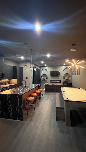 A modern kitchen with a marble island, bar stools, and a pool table in a dimly lit room with recessed lighting and decorative ceiling lights. - META Royal Estate~>THE KAWS HAUS! By Yasmeen (Houston)