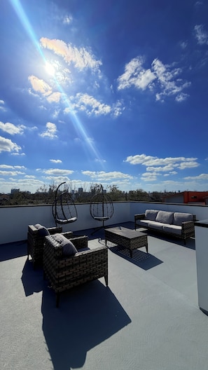A rooftop terrace with wicker furniture, a cushioned sofa, and a view of the city skyline. - META Royal Estate~>THE KAWS HAUS! By Yasmeen (Houston)