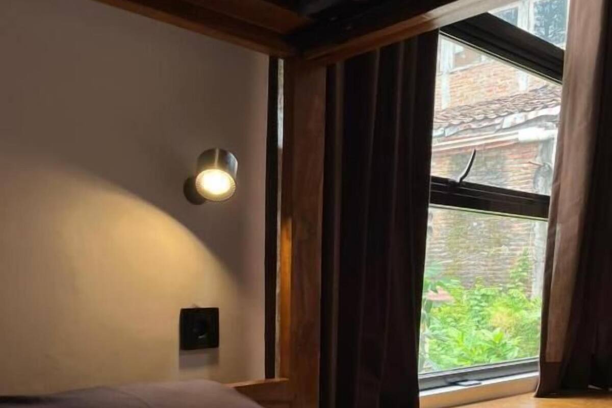 Un dormitorio con cama, ventana con cortinas, luz empotrada en la pared y vistas al exterior.