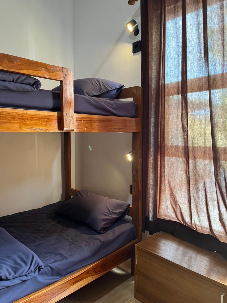 A bunk bed with two levels, a wooden nightstand, and a window with curtains.