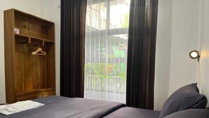 A bedroom with a bed, a wooden wardrobe, a window with curtains, and a wall-mounted light fixture. - Kali Code Home (Yogyakarta)