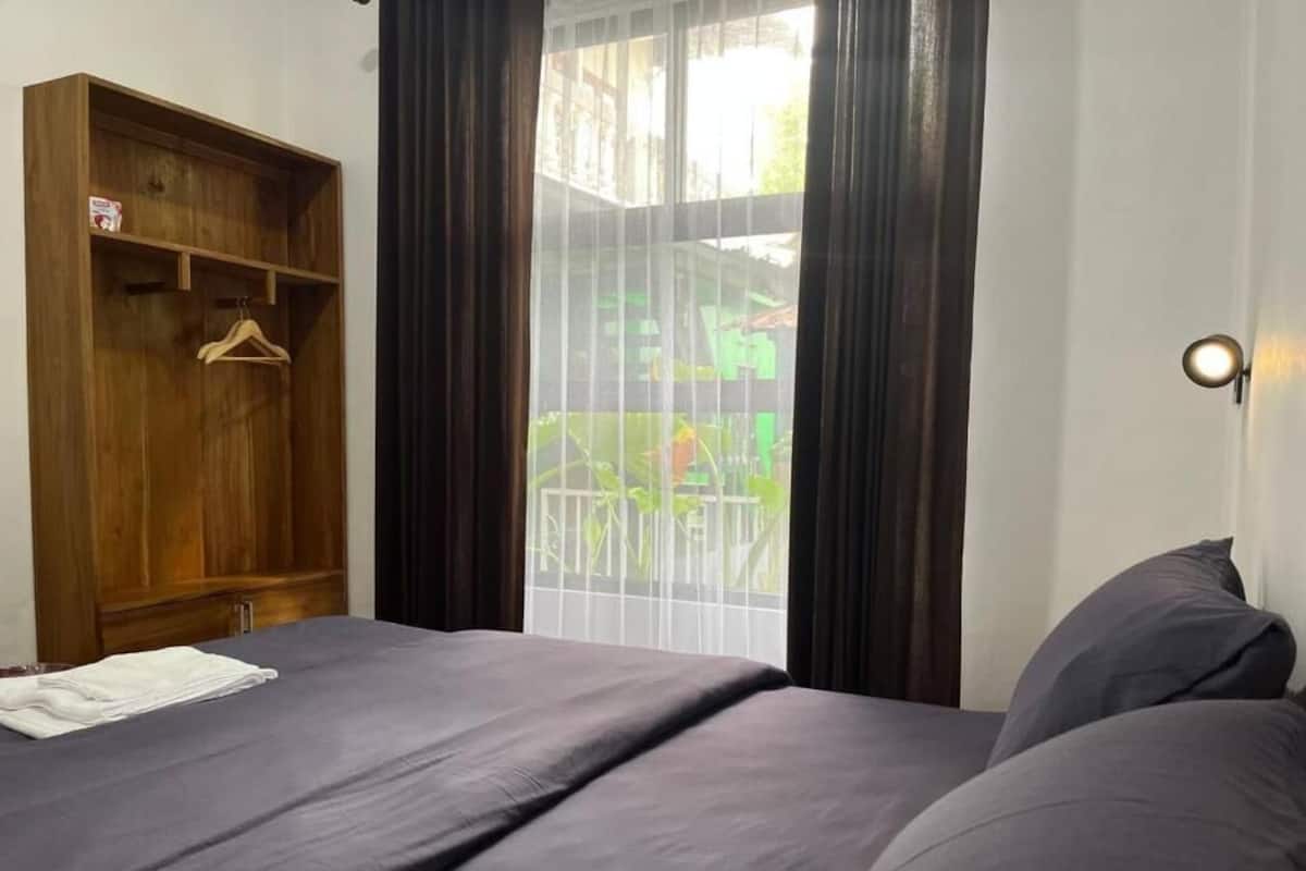 Un dormitorio con cama, un armario de madera, una ventana con cortinas y una lámpara fijada en la pared.