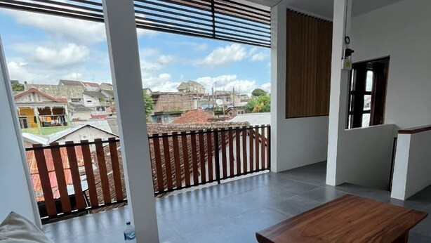 A modern living room with a white sofa, a wooden coffee table, and large windows with a view of a residential area. - Kali Code Home (Yogyakarta)