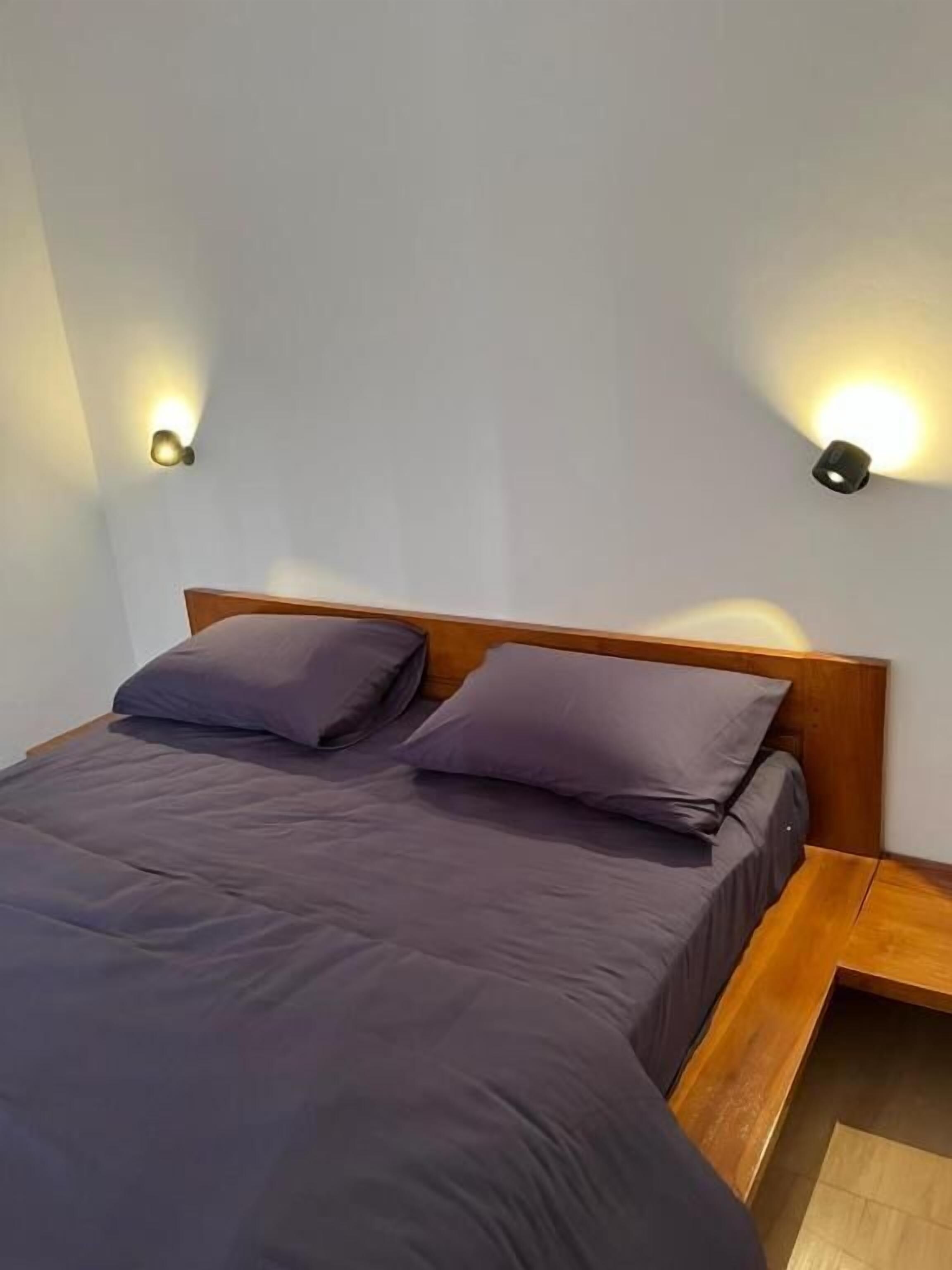 Una cama con cabecera de madera, dos almohadas y dos lámparas fijadas a la pared.