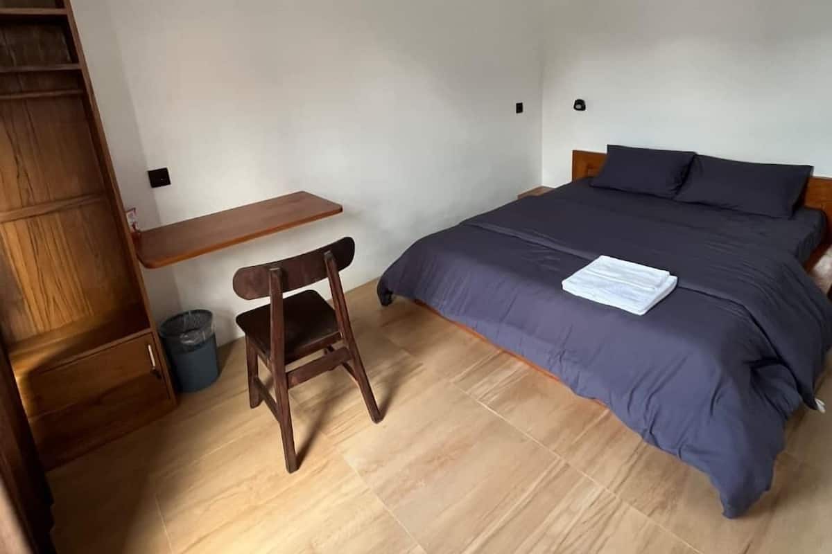 Un dormitorio con una cama, un escritorio de madera, una silla y un cubo de basura pequeño.