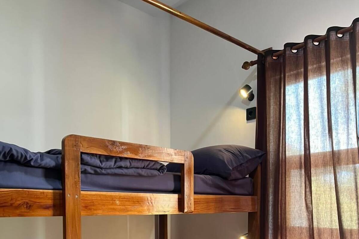 Una cama individual con estructura de madera y ropa de cama oscura, un armario pequeño y una ventana con cortinas.