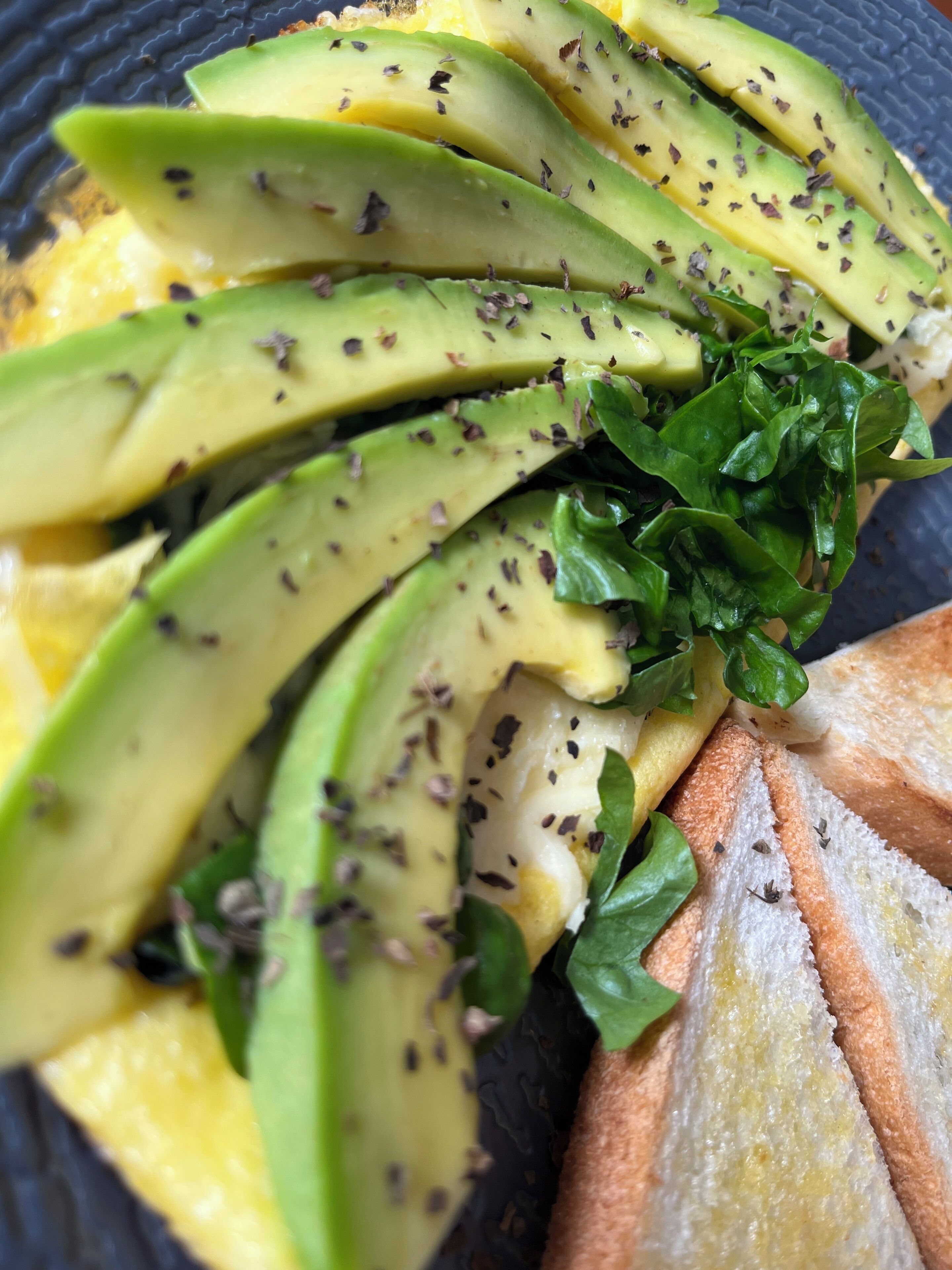 Un plato con aguacate en rodajas, vegetales de hoja verde y pan.