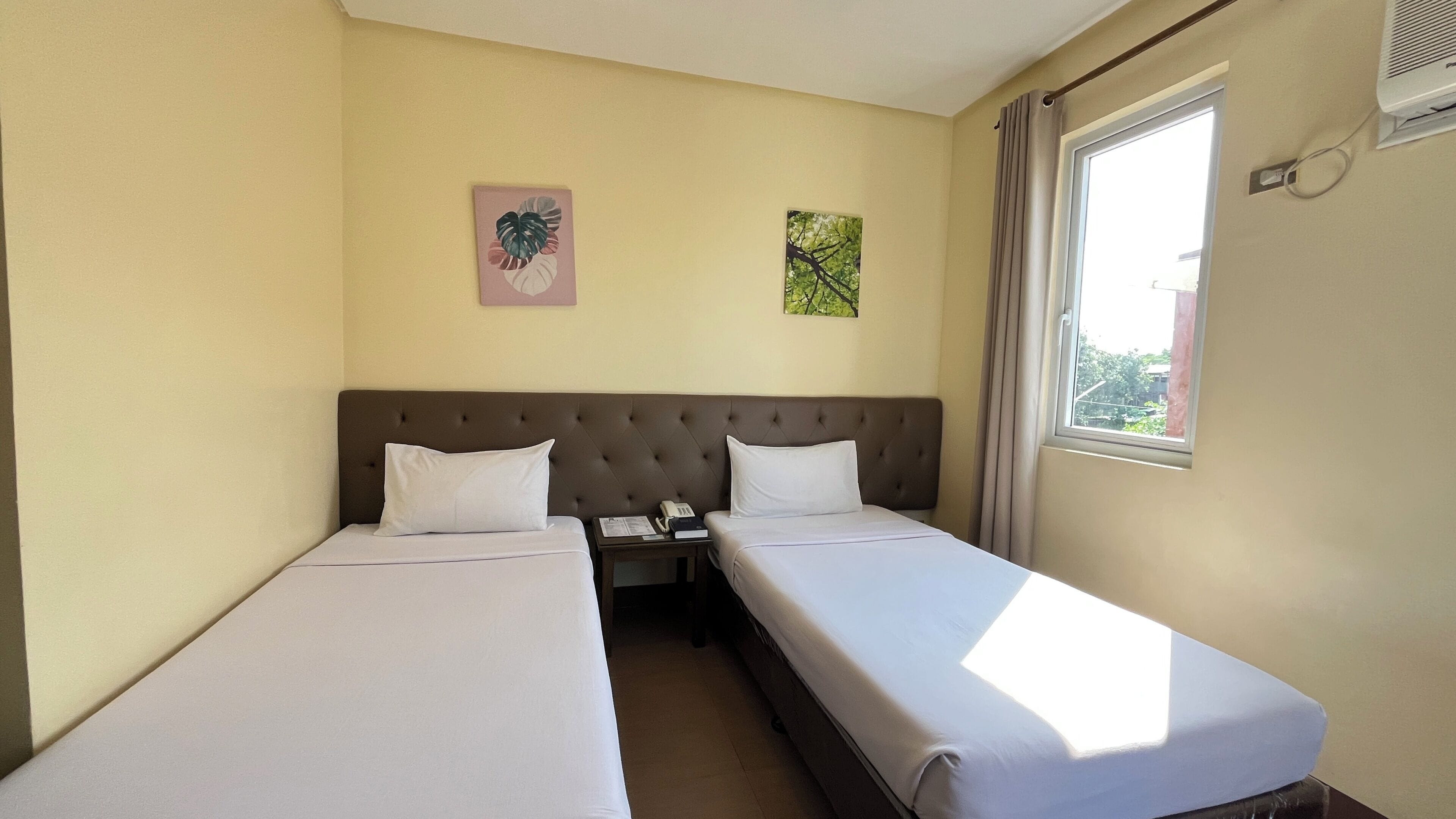 Habitación de hotel con dos camas individuales, una mesita de noche con un teléfono y libros, una ventana con cortinas y dos cuadros decorativos.