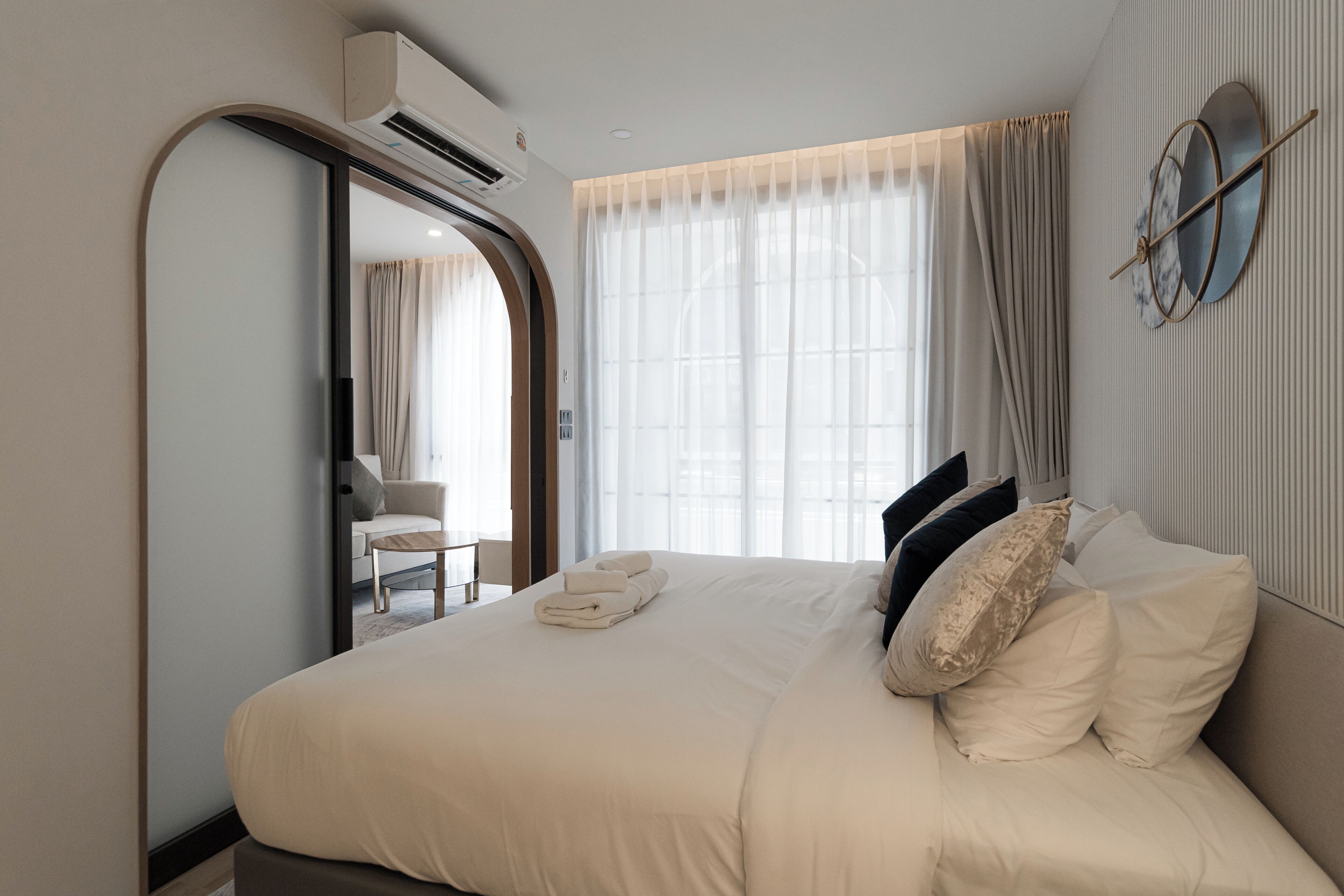 A modern hotel room with a large bed, a round mirror, and a view of the outdoors through sheer curtains.