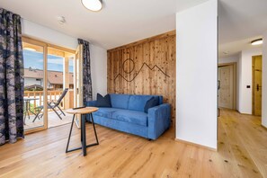 A modern living room with a blue sofa, wooden flooring, and a large window with curtains. - (4) First-floor vacation rental with a balcony /2 (Reit im Winkl)