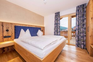 Ein Bett mit Holzrahmen, weißer Bettwäsche und Kopfteil, ein Nachttisch aus Holz sowie ein Blick durch eine Glastür auf einen Balkon.