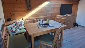 Une chaleureuse cabane en bois avec une table à manger, des chaises, un banc agrémenté de coussins à motifs floraux, un téléviseur et une petite statue posée sur le sol.