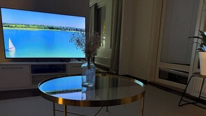 Pyöreä lasipöytä, jossa on metallijalat, maljakko kuivakukilla ja televisio, josta näkyy kaunis järvimaisema.