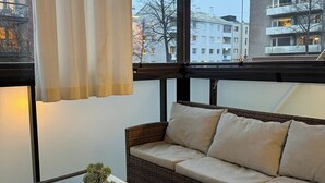 Huoneessa on rottinkisohva, lasinen sohvapöytä, jossa on kasvi, ja suurista ikkunoista avautuu näkymä rakennuksiin.