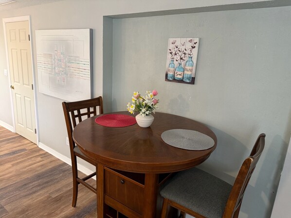 Un coin repas comprenant une table ronde en bois, deux chaises, un vase avec des fleurs et une œuvre d’art accrochée au mur.