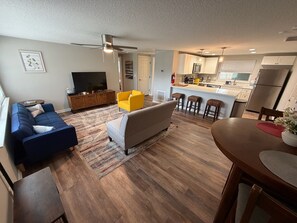 A modern living room with a sofa, armchair, and a dining table with chairs. A kitchen with a bar area is visible in the background.