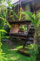 A rustic wooden structure with a bench and a ladder, surrounded by lush greenery and a stone path.