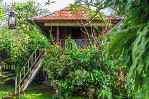 Una casa de madera rústica con techo de tejas, rodeada de abundante vegetación y flores rojas.