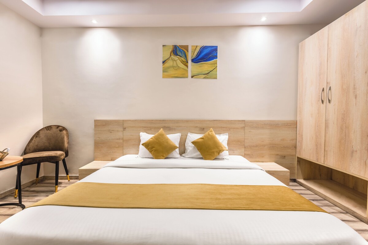 A hotel room with a bed, a wooden wardrobe, a chair, and two paintings on the wall.