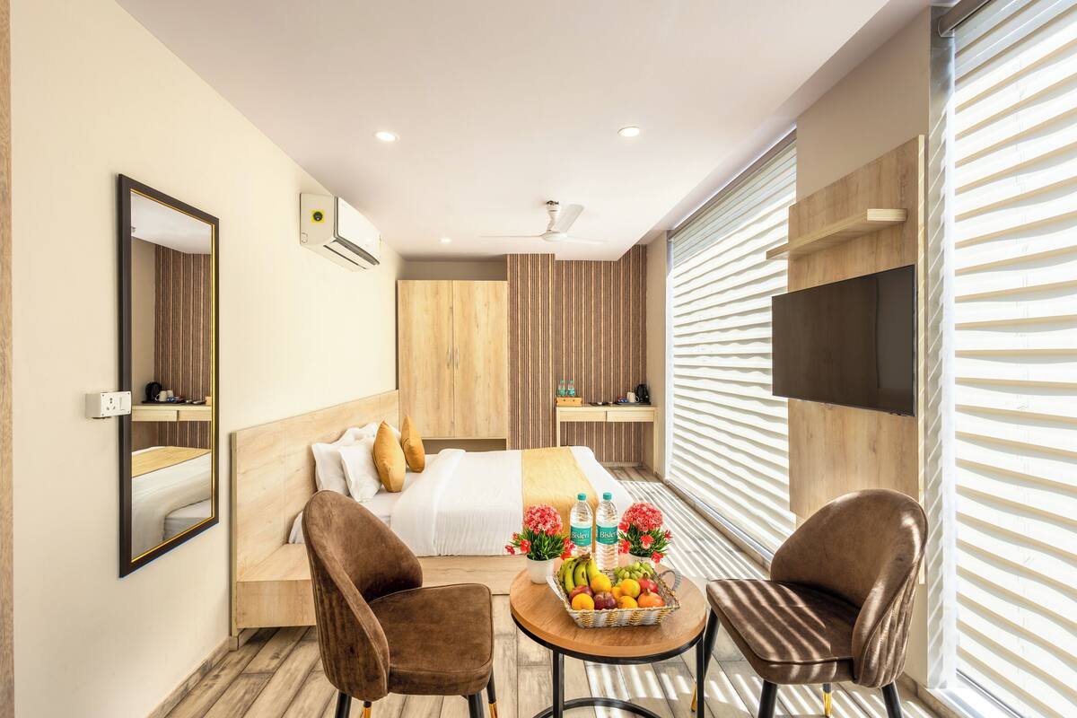 Une chambre d’hôtel moderne, équipée d’un lit, de deux fauteuils, d’une petite table avec des fruits et des fleurs, d’un téléviseur et d’une fenêtre avec des stores.