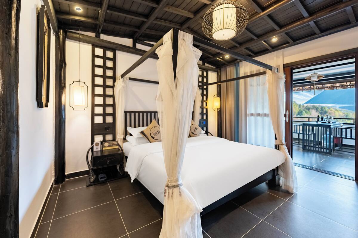 A four-poster bed with a canopy, a bedside table with a lamp, and a view of a balcony with outdoor furniture.