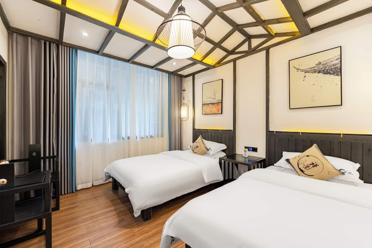 A hotel room with two beds, a desk, a chair, a window with curtains, and artwork on the walls.