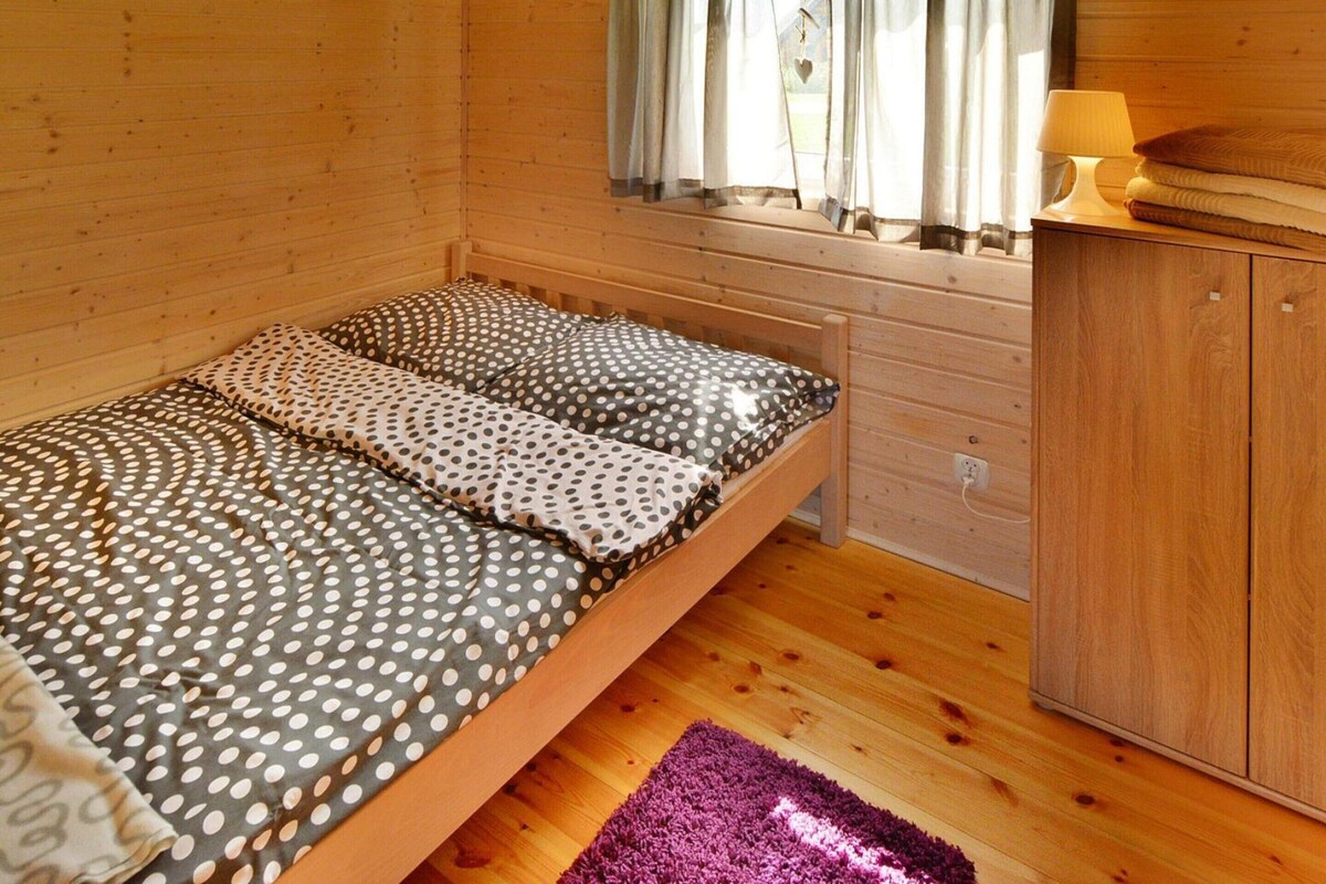 A wooden cabin room with a bed, a window with curtains, a nightstand with a lamp, and a wooden wardrobe.