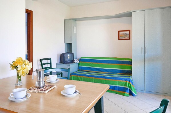 Una habitación compacta con una cama, una mesita con cafetera, tazas y un jarrón de flores.