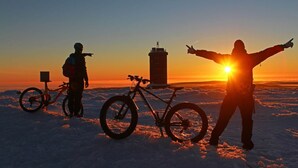 Henkilö seisoo auringonlaskun aikaan lumisessa maisemassa, kädet levällään, ja taustalla näkyy polkupyörä sekä korkea rakennelma.